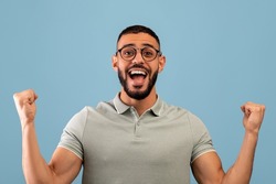 Satisfied happy arab man rejoicing to success and shouting in excitement, standing on blue background. Middle eastern guy celebrate victory, triumph, enjoy success and celebrate win