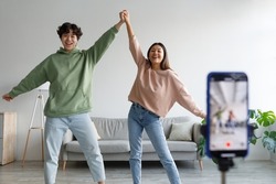 Happy young Asian couple making video on cellphone, dancing in front of camera at home. Lifestyle bloggers creating entertainment content for their vlog, live streaming together