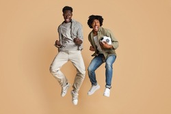 Football Fans. Full Length Shot Of Two Euphoric Black Guys Jumping In Air With Soccer Ball In Hands, Excited African American Friends Emotionally Reacting To Win, Having Fun On Beige Background