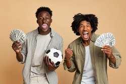 Sport Bets. Portrait Of Excited Black Guys With Football Ball And Money Standing Over Beige Background, Cheerful Soccer Fans Celebrating Success With Dollar Cash, Emotionally Reacting To Win