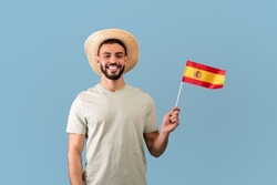Happy arab man wearing casual t-shirt and hat, holding the spanish flag and smiling at camera, standing over blue studio background, copy space