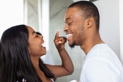 Closeup Shot Of Happy African American Couple Having Fun In Bathroom, Cheerful Black Woman Applying Moisturising Cream On Husband's Face, Playfully Touching His Nose And Laughing Together