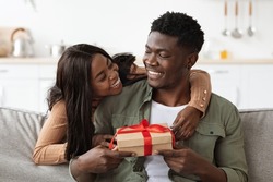 Emotional african american woman hugging lover, giving gift box, making great surprise for St. Valentines Day, home interior, free space. Happy black lady embracing her husband, holding present