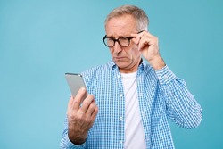 Bad News Concept. Upset Mature Man Holding Smartphone, Looking At Mobile Phone Screen With Worried Expression, Touching Glasses, Sad Adult Male Reading Unplesant Message, Blue Studio Background