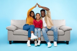 Family Care, Protection And Insurance Concept. Portrait of smiling young African American parents making symbolic roof of hands above their happy daughter sitting on sofa couch at blue studio wall