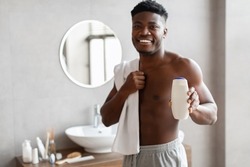 African Guy Showing Shampo Bottle Advertising Male Cosmetic Product Standing In Bathroom Indoor, Wrapped In Towel. Selective Focus On Shower Gel Or Body Lotion In Man's Hand. Shallow Depth