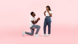 Marry Me. Happy black man holding giving open box with engagement ring to excited girlfriend asking her to be his wife during romantic date standing on one knee, pink studio wall, banner, copy space