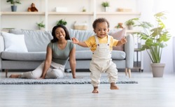 First Steps. Adorable Black Infant Child Walking In Living Room At Home, Cute African American Toddler Boy Stepping On Floor And Looking At Camera, His Proud Mother Smiling On Background, Free Space