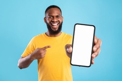 Handsome happy black guy pointing at smartphone with blank screen on blue studio background, mockup for website or new mobile app. Cellphone display template. Space for advertisement