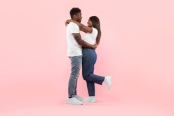 Couple In Love. Portrait of joyful black man hugging his girlfriend, standing together isolated over pink studio background. Casual guy and lady smiling, profile side view, full body length, banner