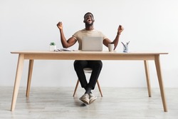 Full length portrait of excited millennial black man gesturing YES in front of laptop, celebrating success or achievement, sitting at desk in home office, copy space. Remote job, online win concept
