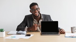 Portrait of happy young black businessman sitting at desk, pointing at laptop computer with blank black screen at home office, mockup. People, technologies and remote work concept