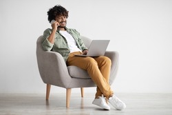 Handsome young indian guy independent contractor working online from home, sitting in comfy armchair, using laptop and having phone conversation with business partner, copy space, full length shot