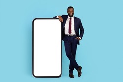 Mobile Offer. Handsome Black Businessman Leaning At Big Blank Mobile Phone, Smiling African American Male Entrepreneur Showing Copy Space For Online Ad Or App Design, Full Length Shot, Mockup