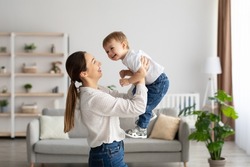Emotional young caucasian mother raising laughing adorable toddler baby son, holding kid pretending flying, having fun and playing together in light living room, side view