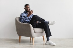 Joyful young black guy sitting in armchair with smartphone, working remotely or video chatting against white wall, free space. Millennial African American male watching movie, having online meeting