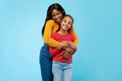 Mother's Love Concept. Portrait of happy young African American mom cuddling her cute tween daughter from the back, standing posing isolated over blue studio background with copy free space