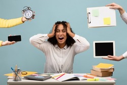 Young African American woman sitting at desk surrounded by hands with alarm clock and gadgets, screaming in despair, feeling exhausted, suffering from burnout. Multitasking, work life balance concept