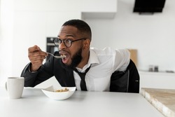 Portrait of busy African American man in glasses having breakfast in kitchen at home, wearing suit while eating cereal, rushing to office, hurry to work in the morning, being late for meeting