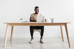 Happy young black man using laptop computer for online work at table in home office, free space. Cool African American guy having remote job, freelancing on web. Modern business concept