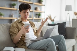 Confused asian man looking at phone screen, sitting on couch with computer on lap in living room, upset frustrated male reading bad news on message, having problem with device or Internet connection