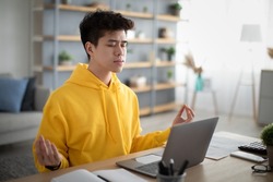 Workplace Stress Management. Calm Asian man meditating with closed eyes in front of laptop pc at home office. Young male freelancer feeling peaceful and balanced, doing yoga sitting at his desk