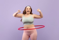 Cheerful millennial european plus size fit lady in sports uniform twirls hula hoop isolated on purple background. Training, motivation, weight loss and body care due covid-19, studio shot, free space