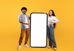 Cool mobile offer. Happy indian couple pointing at big cellphone with white screen for mockup, demonstrating free space for phone application or website design, yellow background