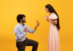 Loving indian man standing on one knee and offering engagement ring to his beloved woman on yellow studio background. Young guy making proposal to sweetheart on Valentine's Day