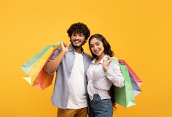Big sale and shopping with pleasure. Happy young indian couple holding many colored packages in hands, posing on yellow background, studio shot, free space