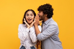 Shock, gossip, share advice. Young indian man whispering to lady on ear, happy woman with open mouth excited and surprised, posing on yellow studio background