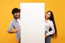 Happy indian couple in love standing by white empty board for advertisement or text over yellow studio background. Excited man and woman standing next to blank placard for ad