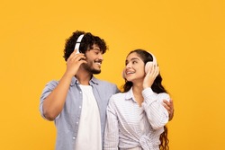 Loving indian couple in headphones listening to music, hugging and looking at each other over yellow studio background. Excited lady enjoying favorite sountracks with her boyfriend