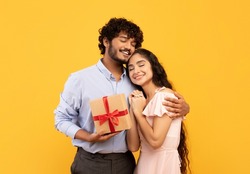 Loving indian man holding wrapped gift box and embracing happy girlfriend, standing over yellow studio background. Romantic young lovers posing with present for Valentine's Day
