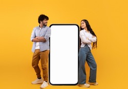 Mobile offer. Millennial indian couple leaning on giant smartphone with mockup, advertising or promoting app or website over yellow studio background
