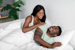 Smiling young pretty african american woman wakes up her husband after night sleep on bed in bedroom. Wake up and good morning, sleep together, love and relationship at home. Couple enjoy free time