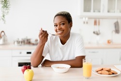 Smiling young attractive black woman eating porridge sits at table with cookies, juice and apples in scandinavian kitchen interior. Good morning, health care, proper nutrition and breakfast at home