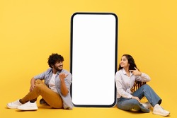 Happy indian couple sitting near big smartphone with blank white screen, demonstrating copy space for app or ad design, posing over yellow background, mockup banner
