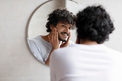 Attractive bearded indian man touching face applying moisturizer on face, standing near mirror in modern bathroom. Male facial skincare routine