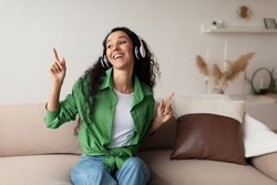 Smiling Middle Eastern Lady Wearing Headphones Listening To Music And Favorite Songs Relaxing Sitting On Couch At Home. Weekend Leisure And Relaxation. Great Playlist Concept