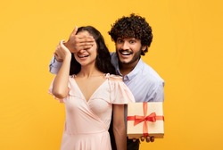 Happy anniversary. Loving indian man covering his girlfriend's eyes, greeting her with wrapped gift box, posing over yellow studio background, celebrating special day
