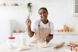 Laughing millennial african american woman in apron prepares dough for baking and raises finger up in light scandinavian kitchen interior. Got idea, cooking pizza, pie at home and facial expression