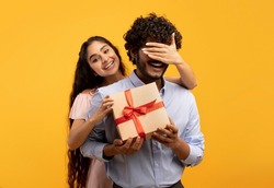 Pretty indian woman covering her boyfriend's eyes, holding wrapped gift box and greeting him with birthday or anniversary, standing over yellow studio background
