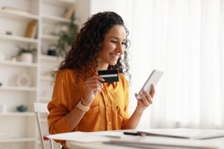 Mobile Shopping. Cheerful Arabic Woman Using Smartphone Shopping Online Holding Credit Card Making Payment Sitting At Desk At Home. Internet Banking Application And E-Commerce. Side View