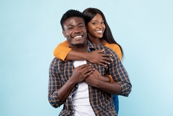 Couple In Love. Portrait of joyful black woman hugging her boyfriend from behind, standing together isolated over blue studio background. Casual guy and lady smiling, posing and looking at camera
