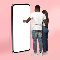 Rear Back View Of Black Couple Using Big Smartphone With Blank White Screen Touching Huge Display Panel With Finger, Guy Higging Lady Standing On Pink Background, Mock Up. Full Body Length