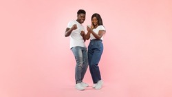 Portrait Of Joyful African American Couple Holding Sharing Smart Phone Shaking Fists In Joy And Excitement, Celebrating Online Win Standing Over Pink Studio Background. Yes, Great News Message Concept