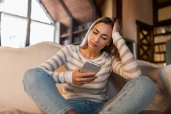 Bad News Concept. Upset Confused Woman Holding Smartphone, Looking At Mobile Phone Screen With Worried Expression, Touching Head, Sad Adult Female Reading Unplesant Message Sitting On Couch