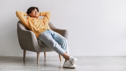 Peaceful young Asian man relaxing in cozy armchair, being lazy, having break against white studio wall, panorama with free space. Handsome millennial guy resting with hands behind head