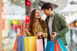 Joyful Couple On Shopping Laughing Holding Colorful Shopper Bags Buying New Clothes In Modern Hypermarket On Weekend. Happy Customers Spending Weekend In Mall. Great Sales Concept